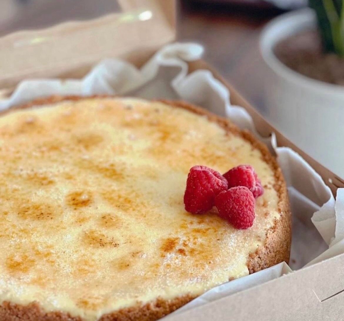 A whole cheesecake with a torched top and fresh raspberries, in a bakery box.