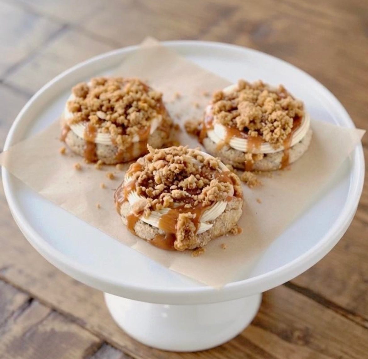 Three crumble-topped cookies with caramel drizzle on a white cake stand.